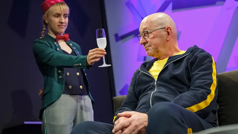 Der Engel Ariel (Tamara Hofer) reicht Simon Notter (Fredi Stettler) ein Glas Champagner.